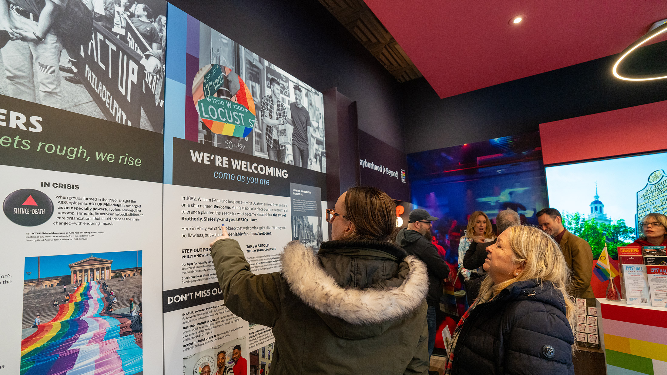 Les visiteurs découvrent les expositions du centre d'accueil Philly Pride, en lisant les panneaux consacrés à l'histoire et à la culture de Philadelphie, tandis que des écrans interactifs et des graphismes colorés sur le thème de la Pride agrémentent cet espace accueillant.