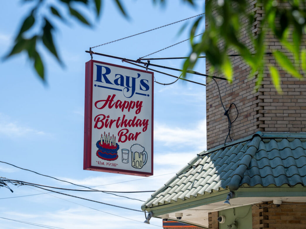 Vintage hanging sign for Ray’s Happy Birthday Bar in Philadelphia, displaying the bar’s name alongside illustrations of a birthday cake, shot glass and beer mug, mounted on a brick building and framed by leafy tree branches against a blue sky.