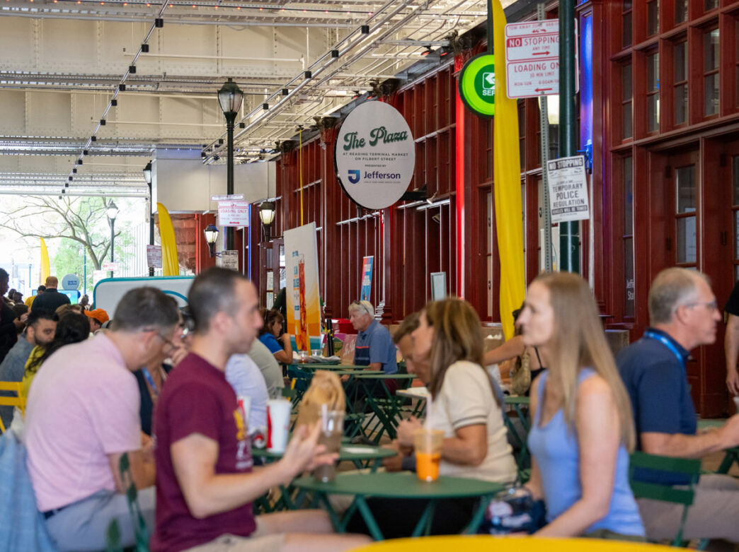 Visitors sit at outdoor tables in The Plaza at Reading Terminal Market, a covered pop-up gathering space along Filbert Street in Philadelphia. People eat, drink, and socialize beneath the market’s elevated structure, with colorful signage and the historic market building lining the bustling scene.