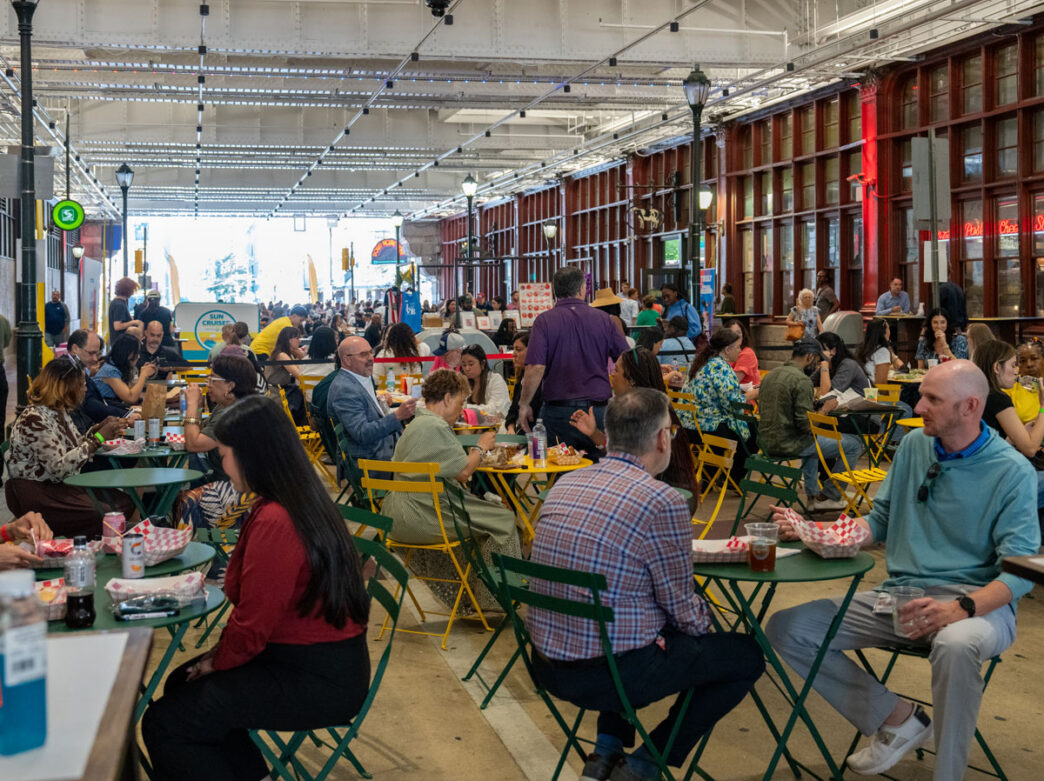 A busy lunchtime crowd sits at green café tables and yellow folding chairs inside The Plaza at Reading Terminal Market in Philadelphia. Diners eat, talk, and socialize beneath the market’s high industrial-style ceiling and exposed steel structure, while food vendors line the perimeter, creating a lively indoor food hall atmosphere.