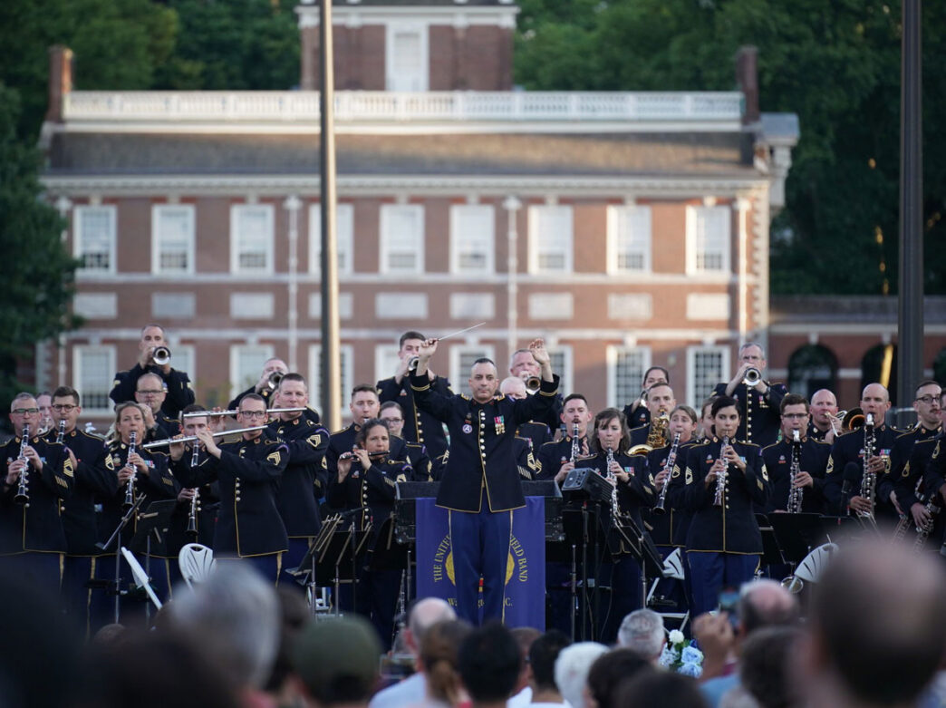 The U.S. Army Field Band performs during the Wawa Welcome America Salute to Service concert in Philadelphia. A conductor leads uniformed musicians playing brass and woodwind instruments on an outdoor stage, with Independence Hall visible in the background and a crowd gathered in the foreground.