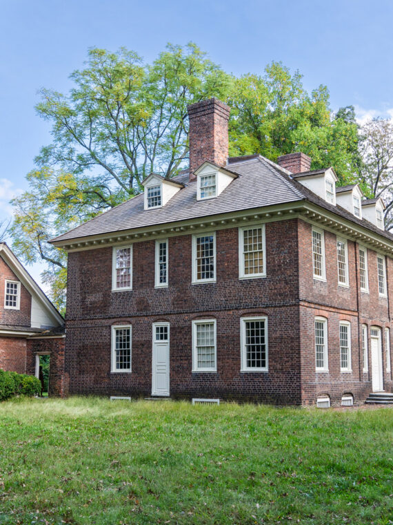 Stenton, a historic 18th-century brick mansion in Philadelphia’s Germantown neighborhood, stands amid a grassy lawn and mature trees. The Georgian-style house features a steep roof, dormer windows, and tall chimneys, highlighting its well-preserved colonial architecture.