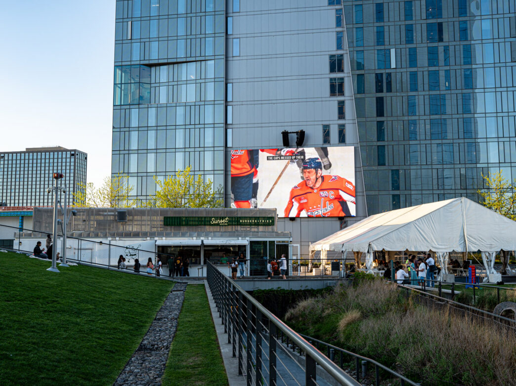 People gather outside at Sunset Social on Cira Green while a hockey game is shown on a large screen against a tall building with a glass facade.