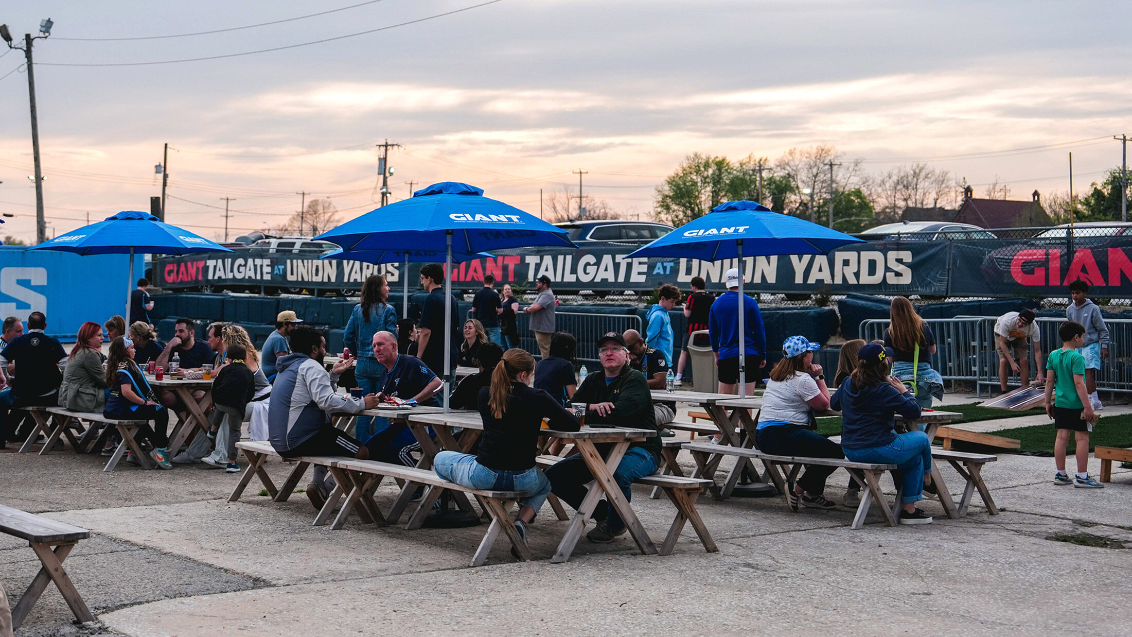 Visitors gather at picnic tables beneath blue umbrellas at Union Yards, an outdoor tailgate and event space adjacent to the Philadelphia Union's Stadium, Subaru Park, in Chester, Pennsylvania.