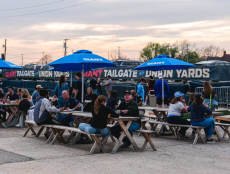 Visitors gather at picnic tables beneath blue umbrellas at Union Yards, an outdoor tailgate and event space adjacent to the Philadelphia Union's Stadium, Subaru Park, in Chester, Pennsylvania.