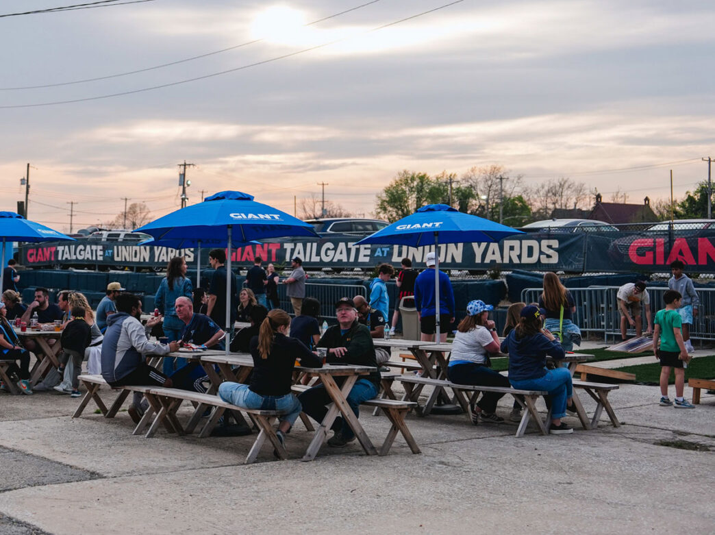 Visitors gather at picnic tables beneath blue umbrellas at Union Yards in Chester, Pennsylvania, enjoying food and conversation in an outdoor event space. Families and friends relax while others play lawn games nearby, with a large Union Yards banner stretching across the background under an evening sky.