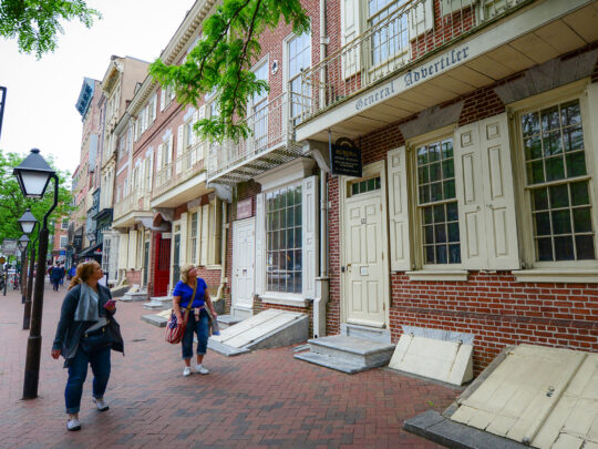 Os visitantes caminham pela Market Street, passando por prédios históricos de tijolos com venezianas na cor creme, incluindo a gráfica de Benjamin Franklin.