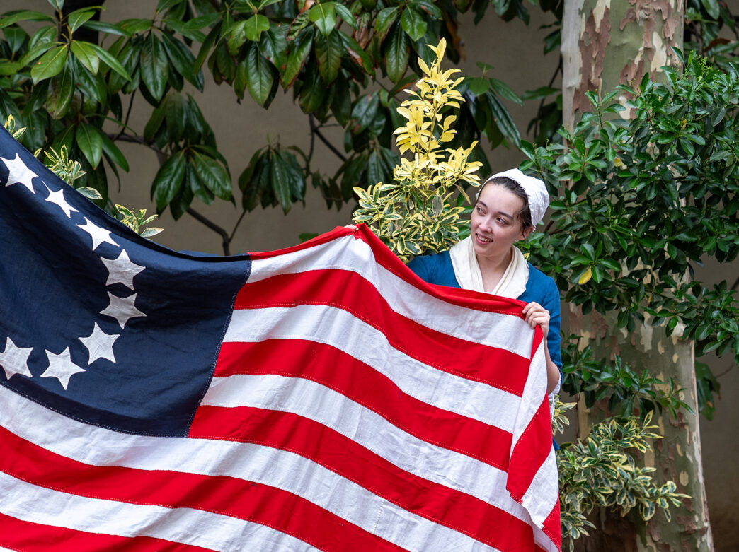 A Betsy Ross reenactor displays a 13-star American flag in front of leafy green trees and bushes.