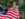 Una intérprete que encarna a Betsy Ross muestra una bandera estadounidense de 13 estrellas frente a frondosos árboles y arbustos.