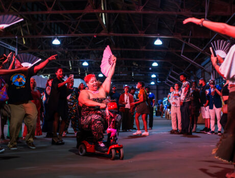 A person riding a mobility scooter through a cheering crowd at a BlackStar Film Festival party, waving an open fan as attendees dance, clap and celebrate together inside a large industrial event space.