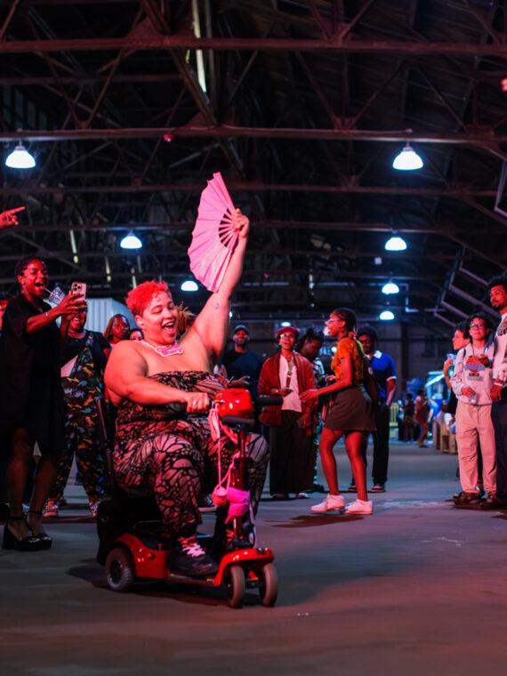 A person riding a mobility scooter through a cheering crowd at a BlackStar Film Festival party, waving an open fan as attendees dance, clap and celebrate together inside a large industrial event space.
