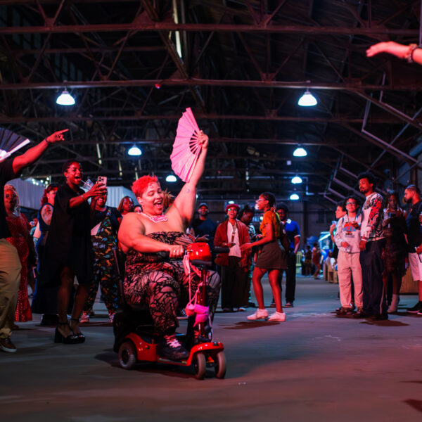 Uma pessoa andando de scooter de mobilidade em meio a uma multidão animada em uma festa do BlackStar Film Festival, acenando com um leque aberto enquanto os participantes dançam, batem palmas e comemoram juntos dentro de um grande espaço industrial destinado a eventos.