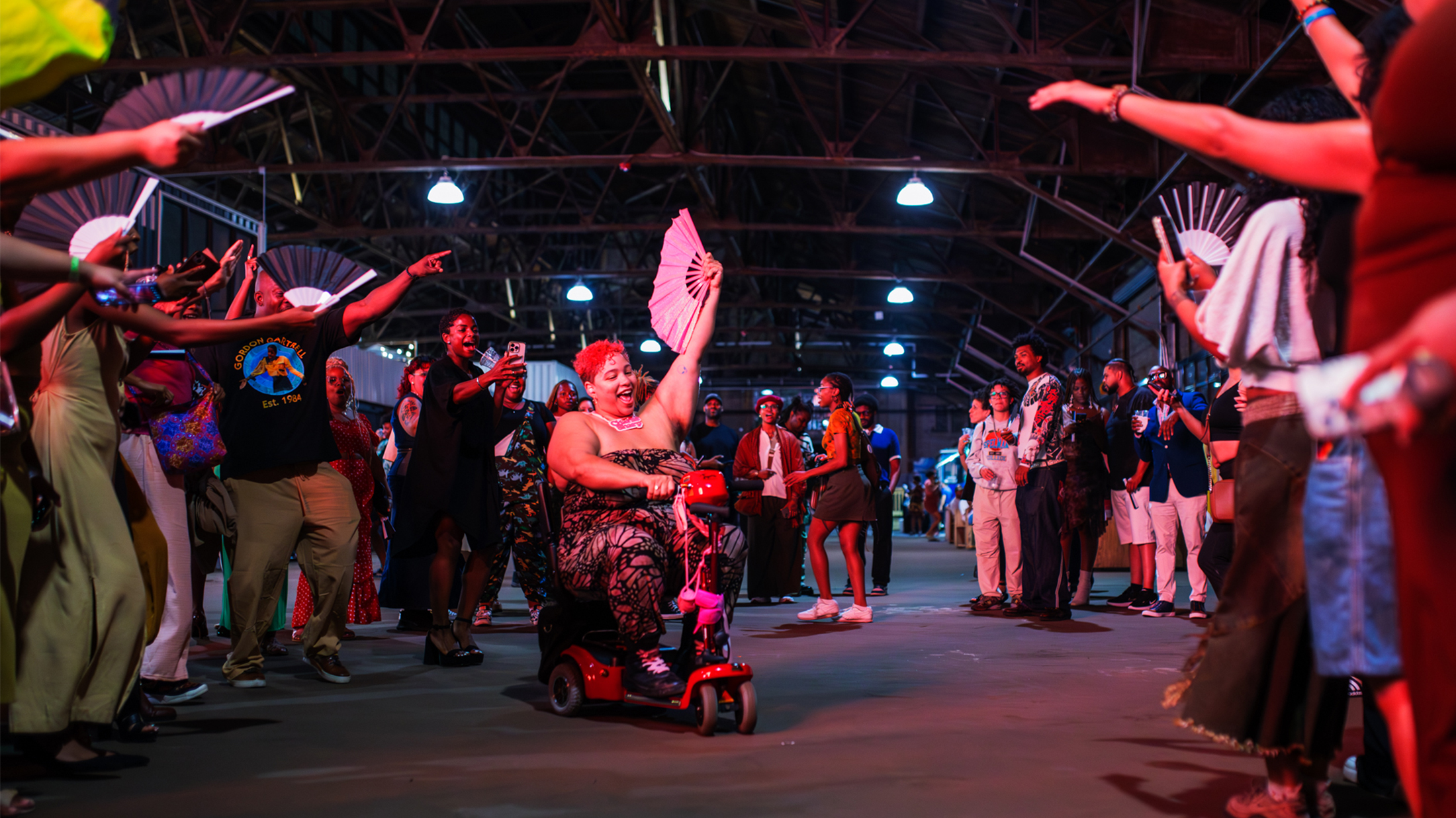 Uma pessoa andando de scooter de mobilidade em meio a uma multidão animada em uma festa do BlackStar Film Festival, acenando com um leque aberto enquanto os participantes dançam, batem palmas e comemoram juntos dentro de um grande espaço industrial destinado a eventos.