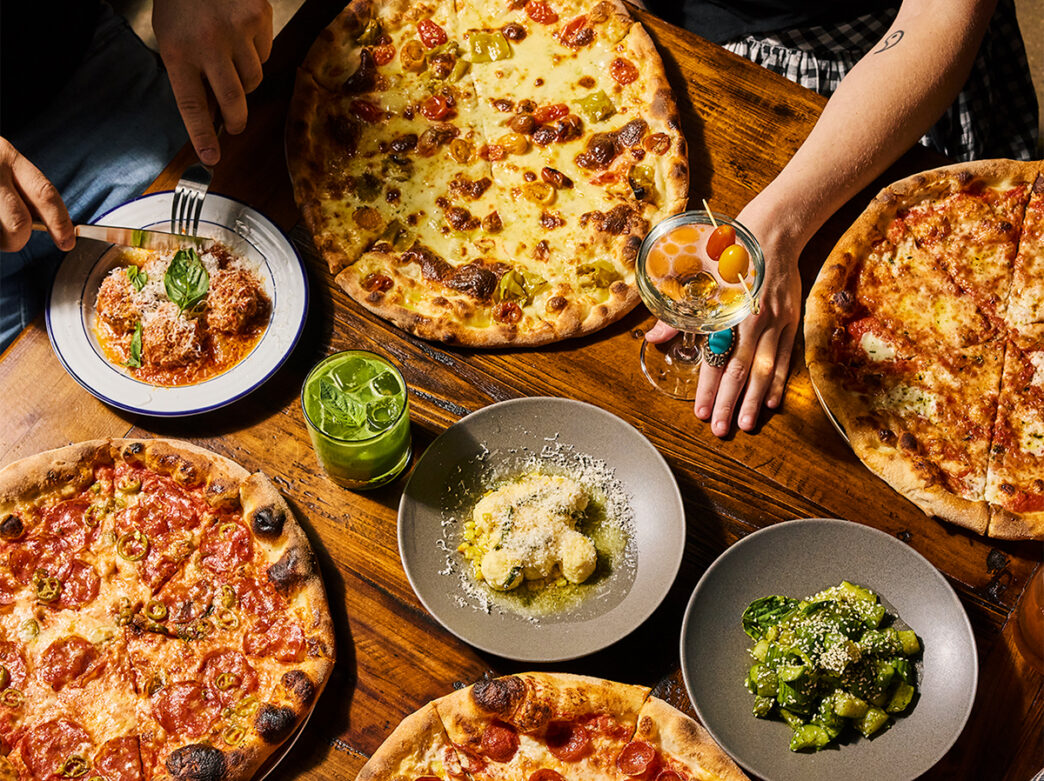 Wood-fired pizzas, pasta dishes, cocktails and small plates arranged on a wooden tabletop as a diner cut into a plate of meatballs.