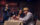 A couple enjoys cocktails at a dining table while a saxophonist and drummer perform live onstage behind them.