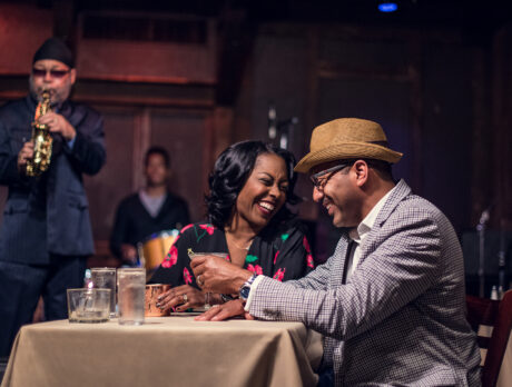 A couple enjoys cocktails at a dining table while a saxophonist and drummer perform live onstage behind them.