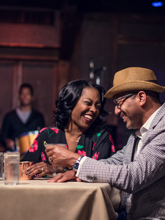 A couple enjoys cocktails at a dining table while a saxophonist and drummer perform live onstage behind them.