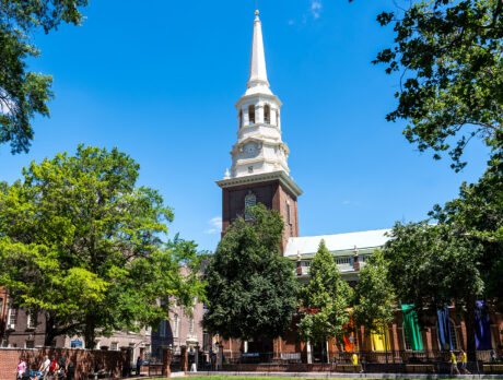 The historic steeple of Christ Church rises above a tree-lined street in Old City, with rainbow banners decorating the exterior of the building.