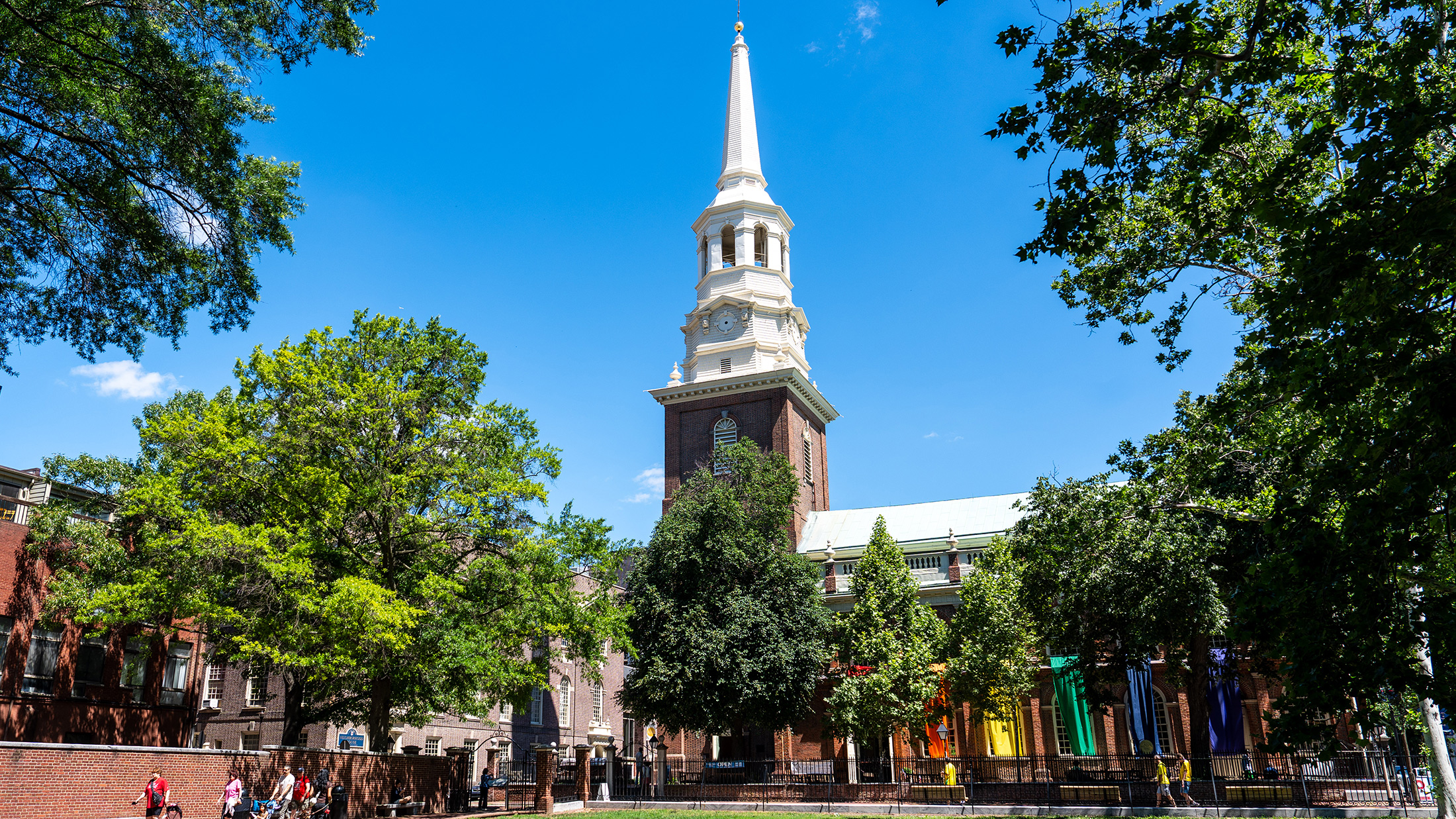 Le clocher historique de Christ Church domine une rue bordée d'arbres dans le quartier Old City, tandis que des bannières aux couleurs de l'arc-en-ciel ornent la façade du bâtiment.