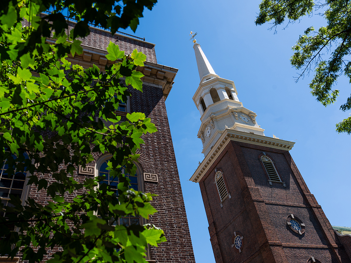 The white steeple of Christ Church rises above leafy trees against a bright blue sky.