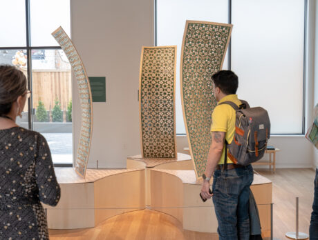 Three people look at a clay installation on display at the Clay Studio in Philadelphia. The installation includes 3 curved panels on elevated platforms.