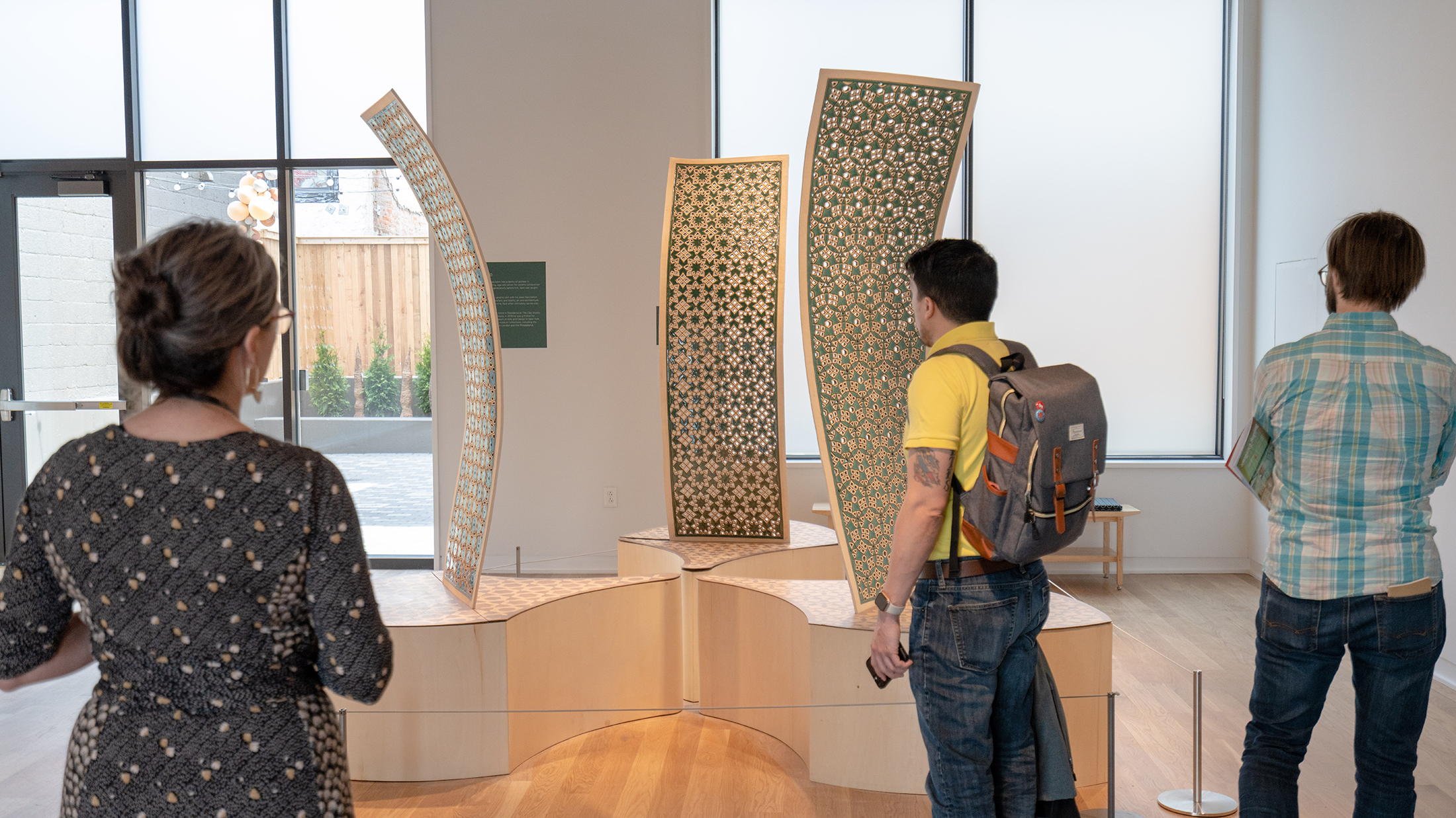 Three people look at a clay installation on display at the Clay Studio in Philadelphia. The installation includes 3 curved panels on elevated platforms.