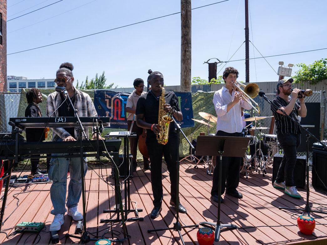 Un ensemble de jazz se produit en direct sur une scène en plein air lors de l'événement marquant le lancement de « Coltrane 100 », composé d'un claviériste, d'un saxophoniste, d'un tromboniste, d'un bassiste et d'un batteur.