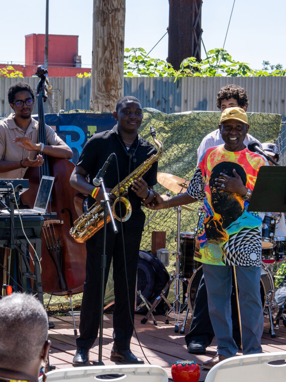Members of a jazz ensemble clap while the saxophonist receives recognition while on on outdoor stage at the Coltrane 100 launch event.
