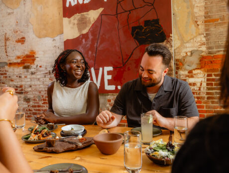Guests sit around a wooden table at Honeysuckle Restaurant, sharing plates of food and cocktails while laughing together beneath exposed brick walls and a large piece of artwork.