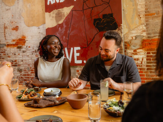 Guests sit around a wooden table at Honeysuckle Restaurant, sharing plates of food and cocktails while laughing together beneath exposed brick walls and a large piece of artwork.