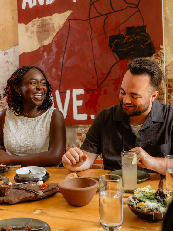 In restaurant Honeysuckle zitten de gasten rond een houten tafel, waar ze gerechten en cocktails delen terwijl ze samen lachen onder de bakstenen muren en een groot kunstwerk.