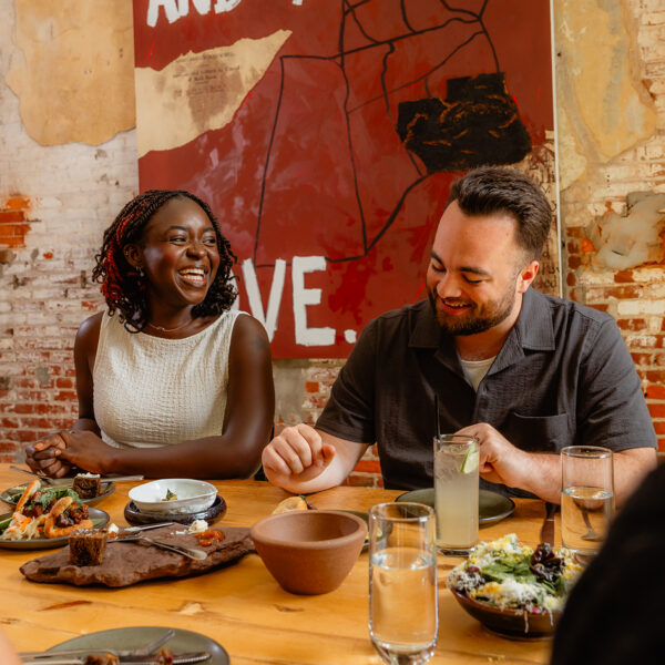 Los comensales se sientan alrededor de una mesa de madera en el restaurante Honeysuckle, compartiendo platos y cócteles mientras se ríen juntos bajo paredes de ladrillo a la vista y una gran obra de arte.