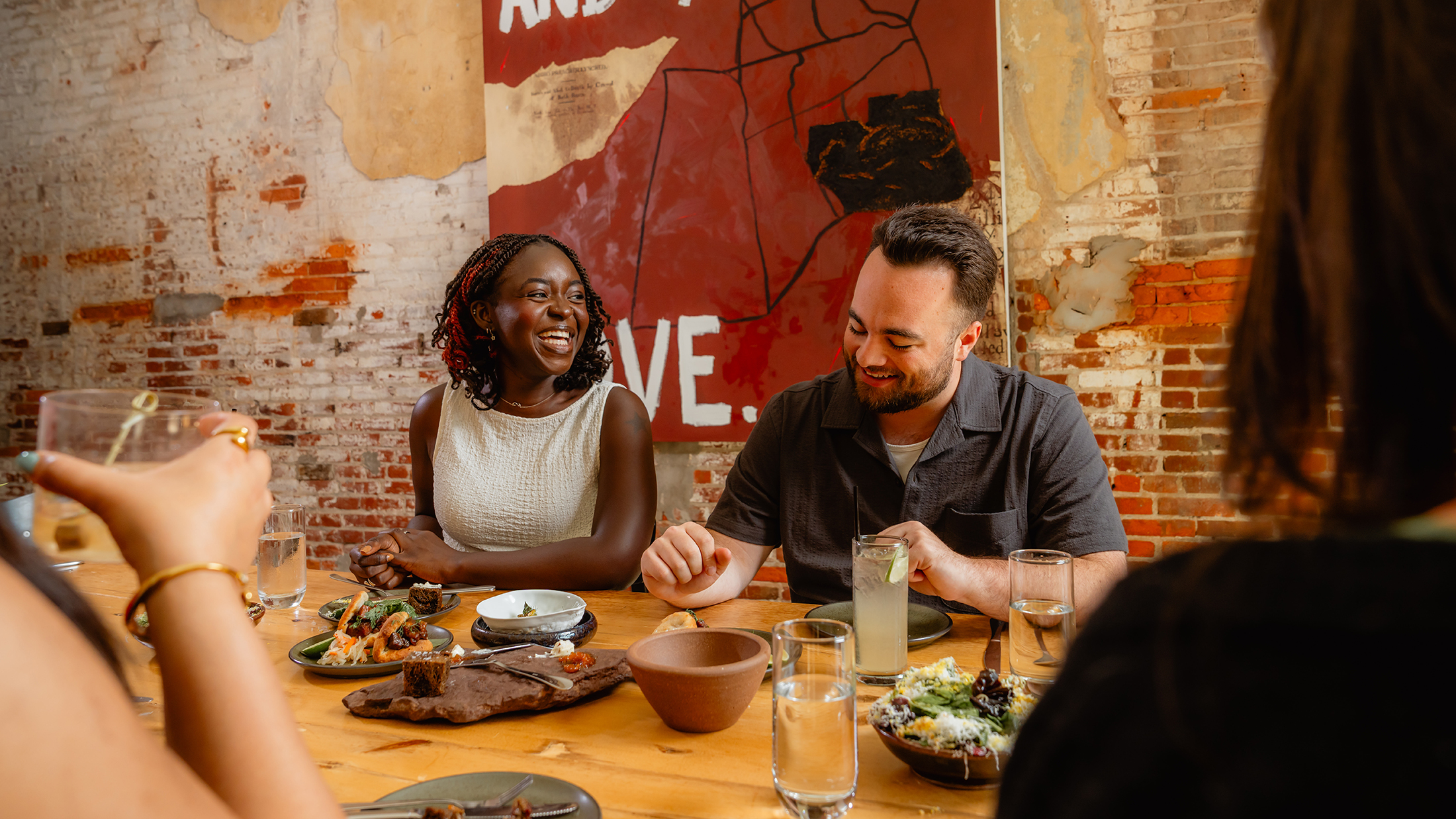 Guests sit around a wooden table at Honeysuckle Restaurant, sharing plates of food and cocktails while laughing together beneath exposed brick walls and a large piece of artwork.