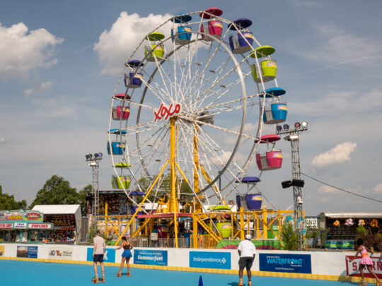 Visitors roller skate on a blue skating rink beneath a colorful Ferris wheel on a sunny summer day.