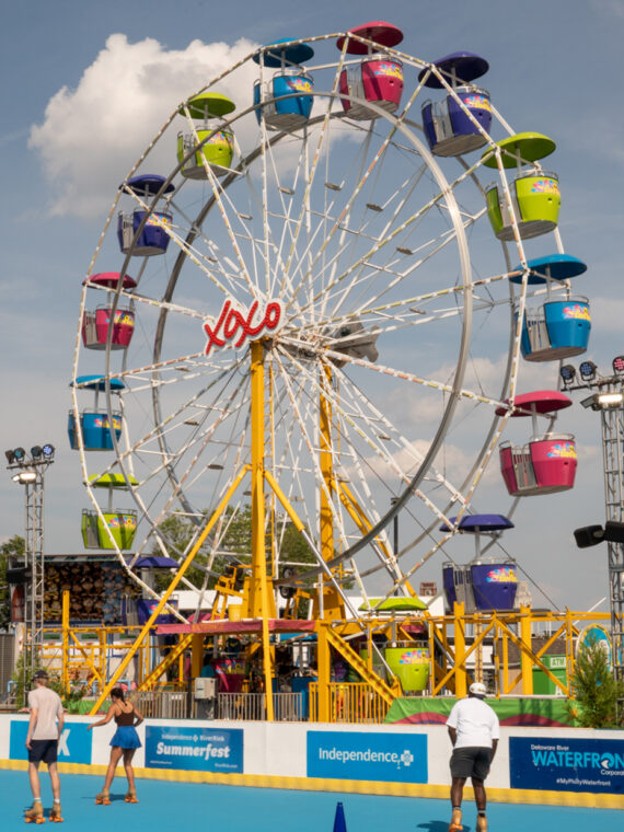 Visitors roller skate on a blue skating rink beneath a colorful Ferris wheel on a sunny summer day.
