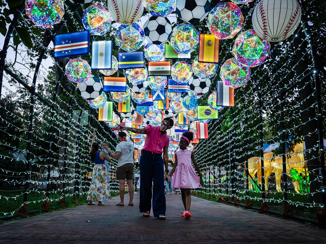 An adult and child walk hand in hand through a tunnel of illuminated soccer ball and international flag lanterns at the Chinese Lantern Festival.