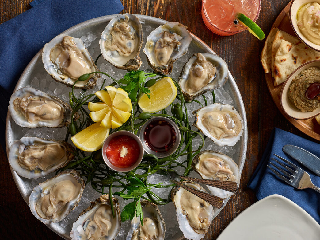 A chilled platter of fresh oysters on the half shell is served over ice with lemon wedges and cocktail sauce on a wooden table with cocktails and pita.