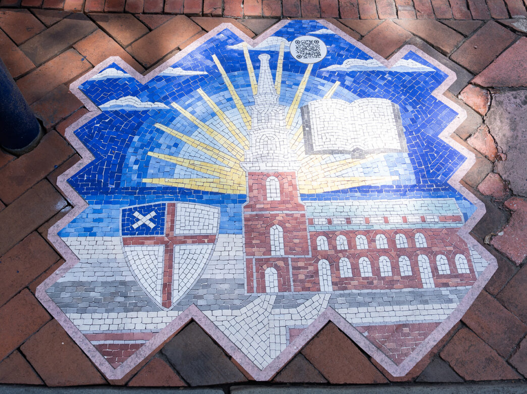 Un primer plano de uno de los marcadores de mosaico hechos a mano del «Camino hacia la Democracia» muestra un edificio histórico, un libro, un sol y un escudo pintados sobre una acera de ladrillos.
