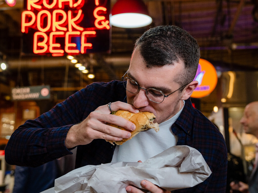 A man takes a bite of a roast pork sandwich wrapped in paper at DiNic’s, with a glowing neon “Roast Pork & Beef” sign behind him.