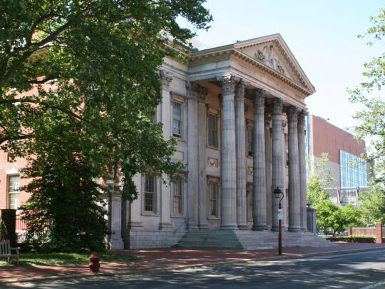 The grand front of the First Bank of the United States shows its stately row of columns and symmetrical stone architecture under a bright blue sky.