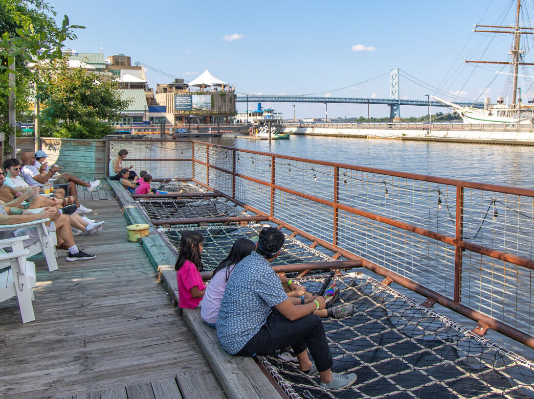 Les visiteurs se détendent dans des chaises Adirondack blanches et des hamacs en filet de chargement, face à la rivière Delaware, avec le Benjamin Franklin Bridge et le voilier historique en arrière-plan.