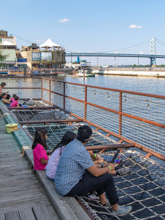 Los visitantes se relajan en sillas Adirondack blancas y hamacas de red de carga con vista al río Delaware, con el Benjamin Franklin Bridge y el velero histórico al fondo.
