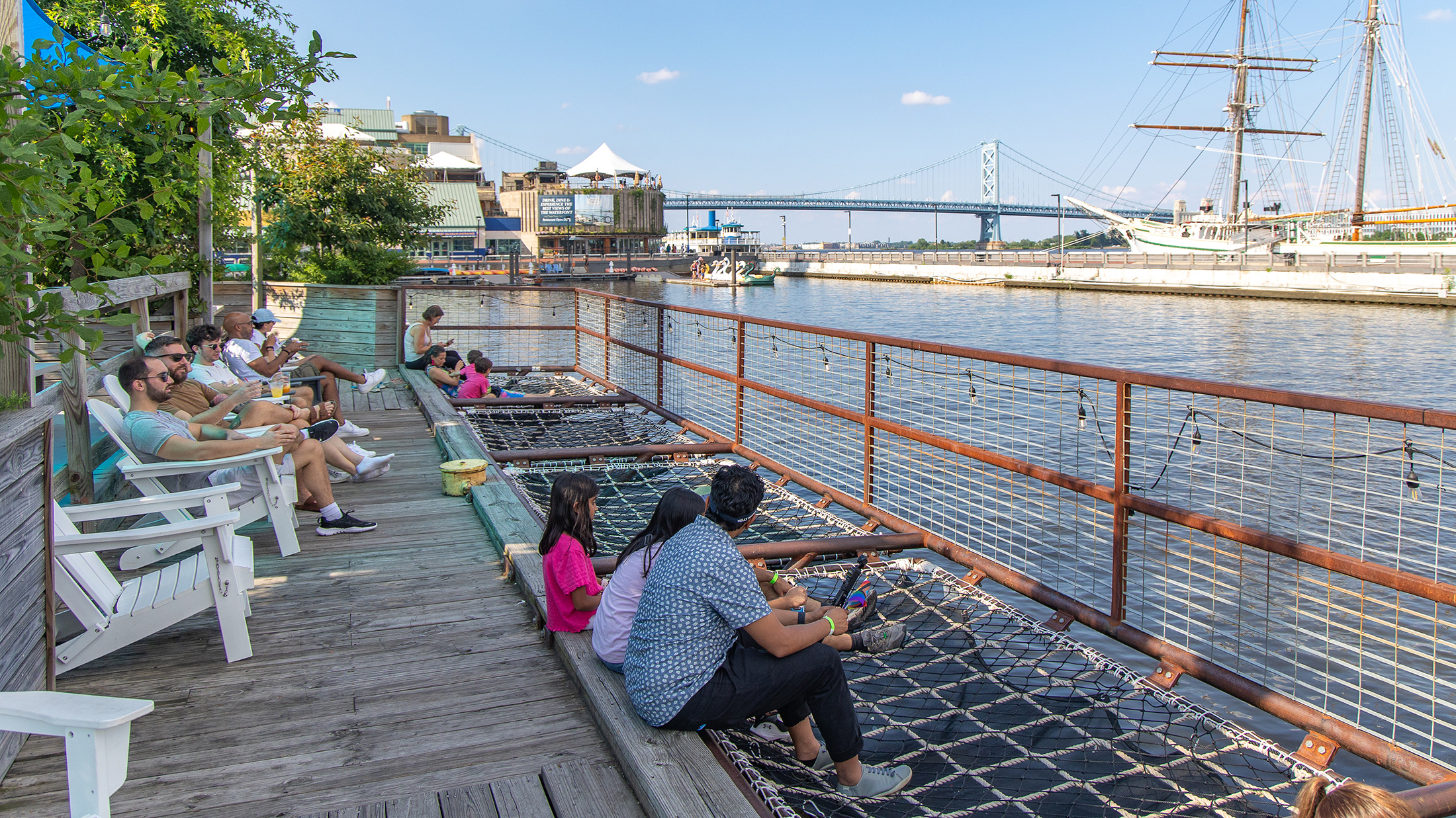 Visitors relax in white Adirondack chairs and cargo net hammocks overlooking the Delaware River, with the Benjamin Franklin Bridge and historic tall ship in the background.