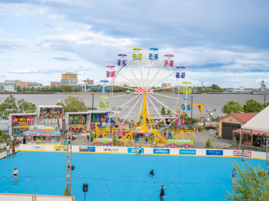 A wide view of Independence Blue Cross RiverRink Summerfest along the Delaware River Waterfront, featuring a blue roller skating rink, colorful Ferris wheel, arcade games and food stands.