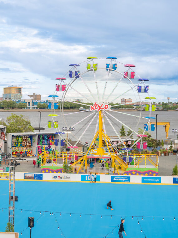 A wide view of Independence Blue Cross RiverRink Summerfest along the Delaware River Waterfront, featuring a blue roller skating rink, colorful Ferris wheel, arcade games and food stands.