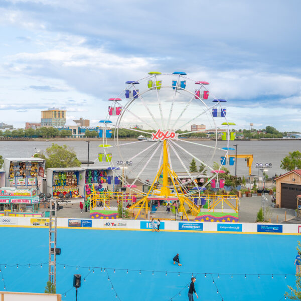 Une vue d'ensemble du festival « Independence Blue Cross RiverRink Summerfest » organisé le long du Delaware River Waterfront, avec notamment une piste de roller bleue, une grande roue colorée, des jeux d'arcade et des stands de restauration.