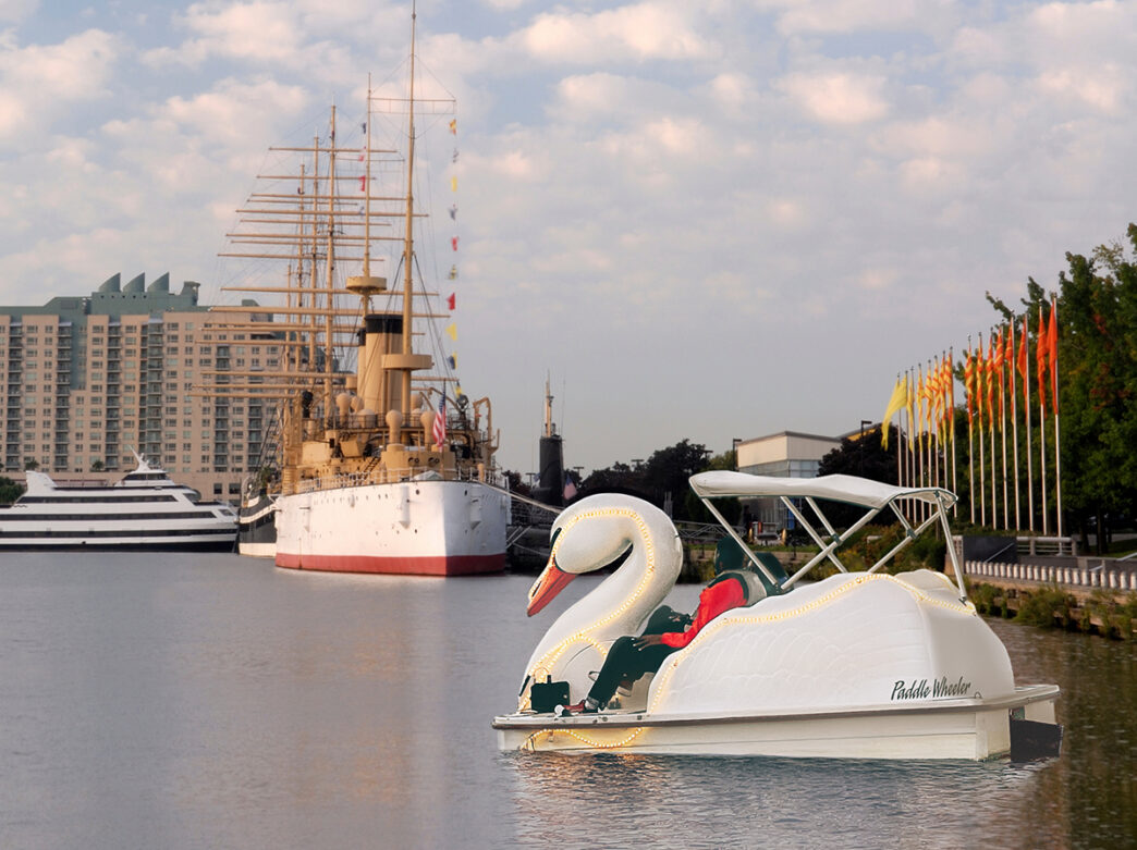 Una pareja pedalea en un bote con forma de cisne luminoso por el río Delaware, con el crucero histórico Olympia visible al fondo.