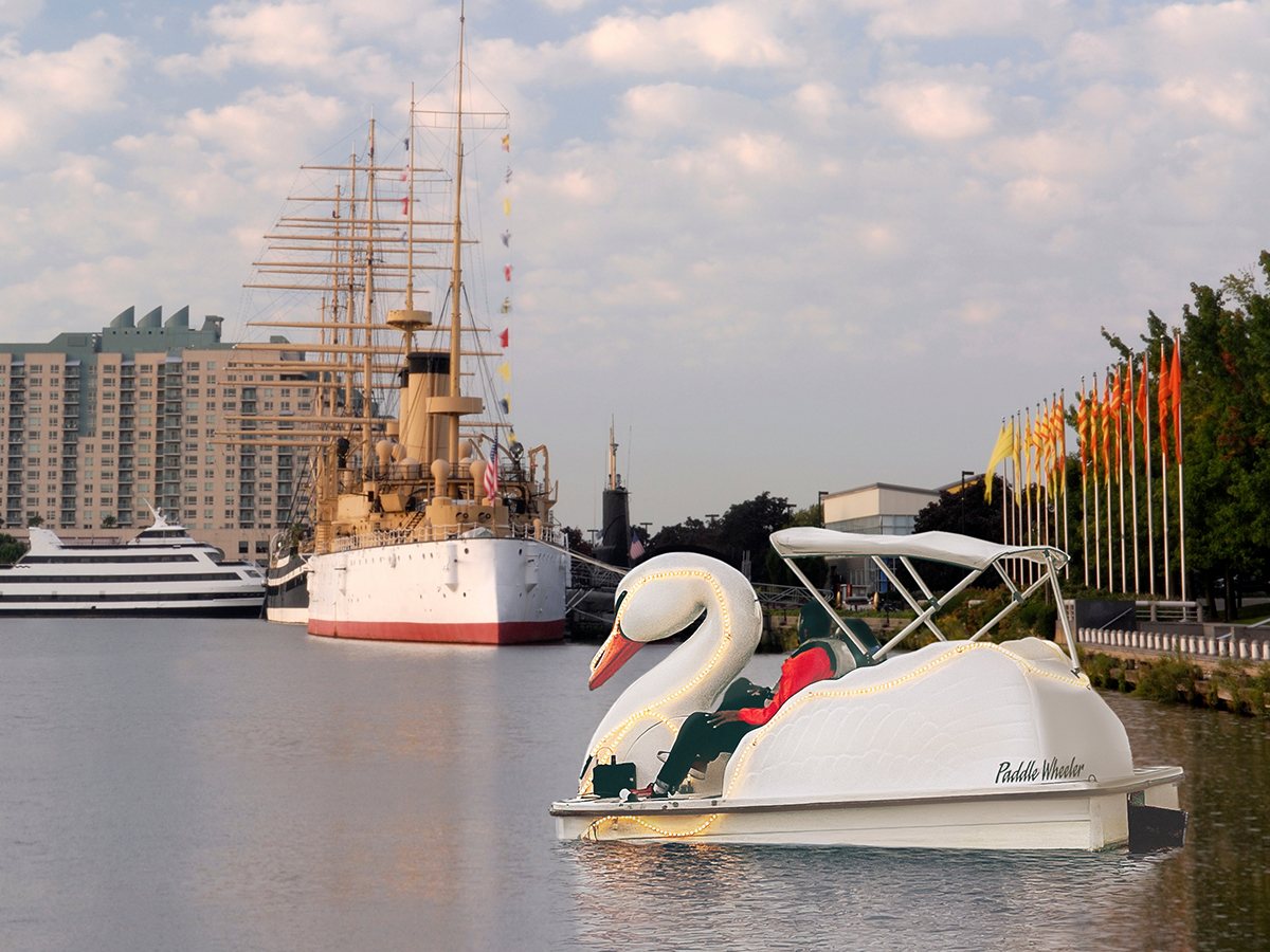 A couple pedals a glowing swan boat along the Delaware River with the historic cruiser Olympia visible in the background.