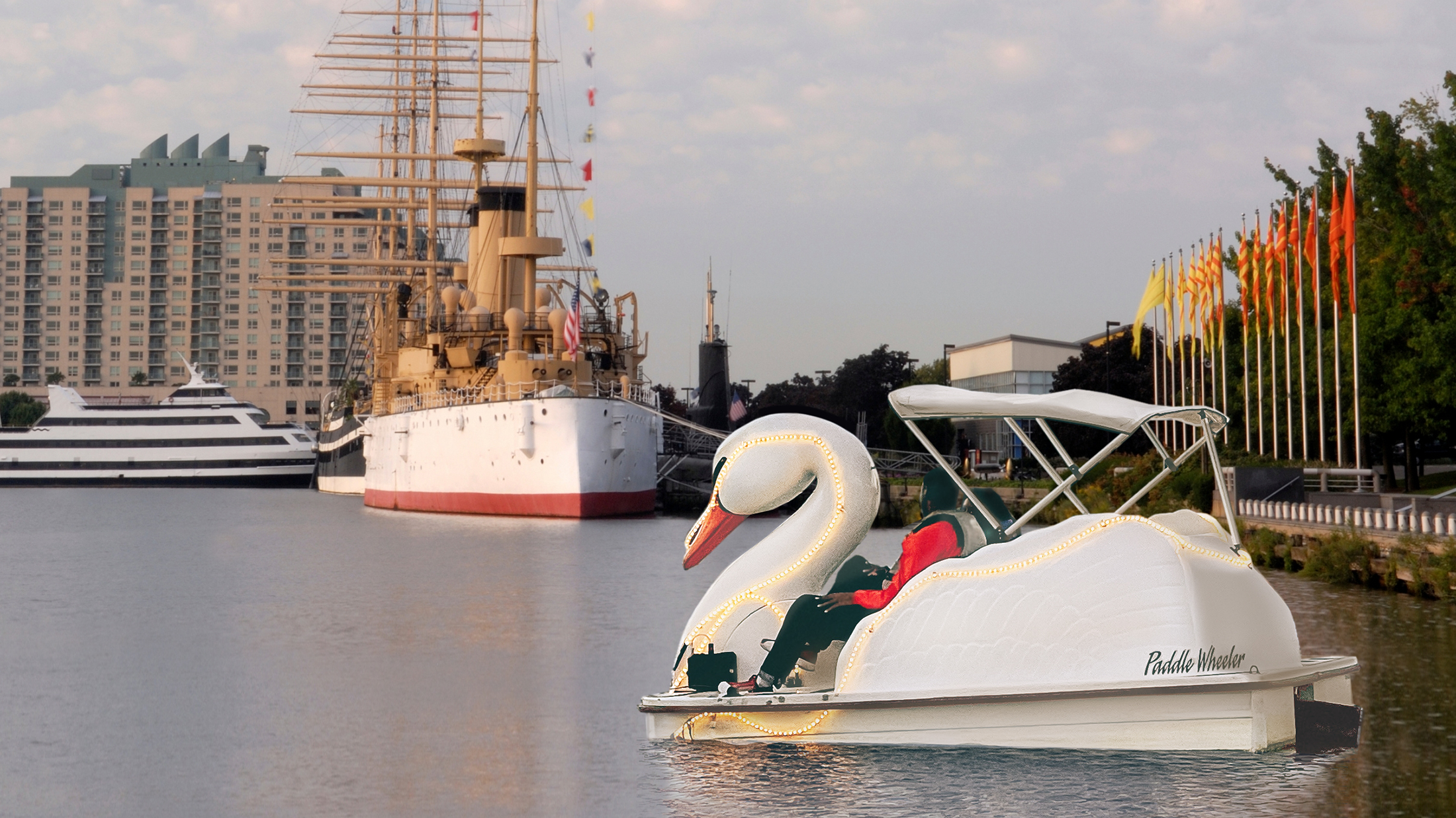 Un couple pédale sur un bateau en forme de cygne lumineux le long du fleuve Delaware, avec le croiseur historique Olympia visible en arrière-plan.