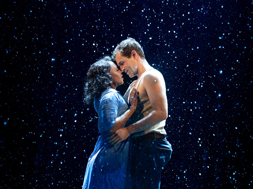 Two performers share an emotional embrace beneath a theatrical rainfall during a performance of The Notebook musical.